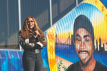 A Black woman, Gina Crowder, with long brass wavy hair stands confidently with a mic, wearing sunglasses and a camo print jacket. A colorful mural of Oscar Grant in front of the Bay Bridge is in the background. 