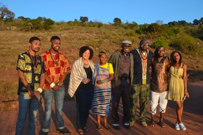 A group of people in an outside setting, with some wearing brightly patterned cultural clothing. There are both younger and older Black individuals, with an expansive field and trees in the background. 