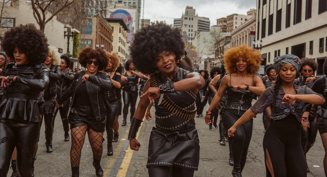 The image shows a person with a large afro and studded leather attire dancing in a street parade, surrounded by others in similar black outfits, exuding confidence and celebration.